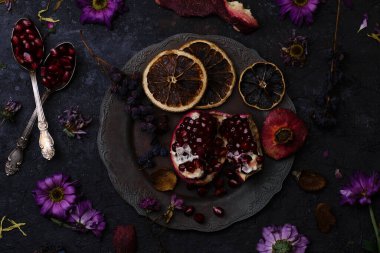 Eski bir tabakta olgun nar, solmuş çiçekler, fiijoa kabuğu, kuru meyveler ve gümüş kaşıklarda tohumlar.