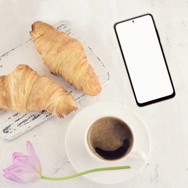 Provence tarzı ahşap bir tahtanın üzerinde taze iki kruvasan ve açık gri arka planda bir fincan sıcak siyah espresso ile modern bir akıllı telefonun kopyası. Üst manzara, düz uzanma