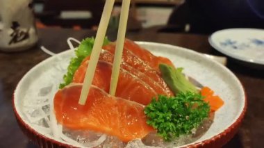 Elleri somon balığını tutmak için çubukları tutuyordu. Somon balığı eski bir masada, üzerinde fotokopi alanı olan siyah bir taş tabağa yerleştirilmişti..
