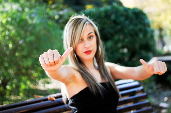 Menina bonita sentado em um banco no parque mostrando sinal de polegar para cima — Fotografia de Stock