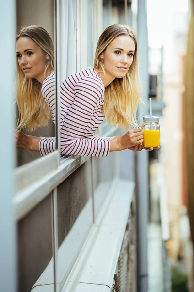 Mujer joven bebiendo un vaso de jugo de naranja natural, asomándose por la ventana de su casa. — Foto de Stock