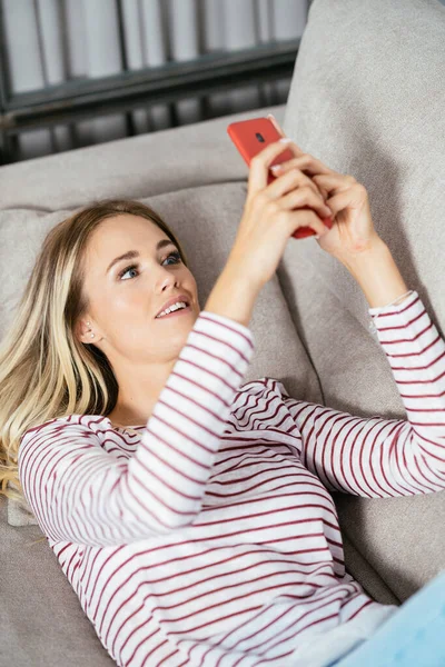 Mujer sonriente usando su teléfono inteligente tumbado en el sofá. —  Fotos de Stock