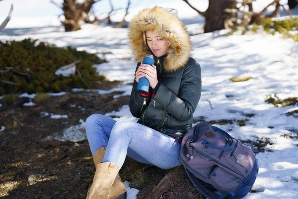 Femme buvant quelque chose de chaud dans une bouteille thermos en métal assise sur un rocher dans les montagnes enneigées. — Photo