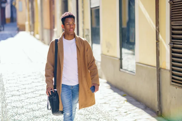 Mladý černoch kráčí po ulici s aktovkou a smartphonem. — Stock fotografie