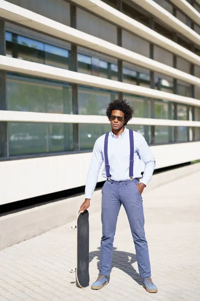 Nero maschio lavoratore in piedi accanto a un ufficio edificio con un skateboard. — Foto Stock