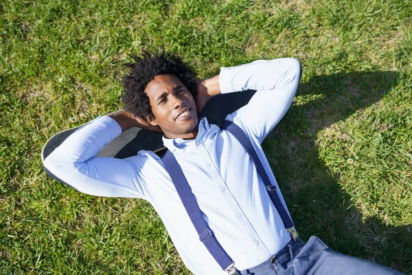Černý podnikatel odpočívající na trávě se svým skateboardem — Stock fotografie