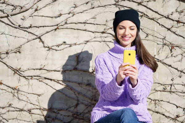 Boldog fiatal nő mosolyog ül a padon a szabadban egy okostelefon segítségével. — Stock Fotó
