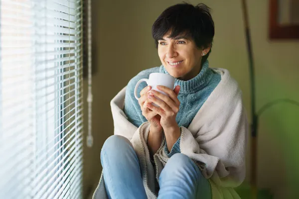 Šťastná žena středního věku se usmívá u okna, popíjí šálek kávy. — Stock fotografie