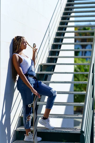 Ung svart kvinna med afro flätor på en urban trappa, bär en skateboard. — Stockfoto