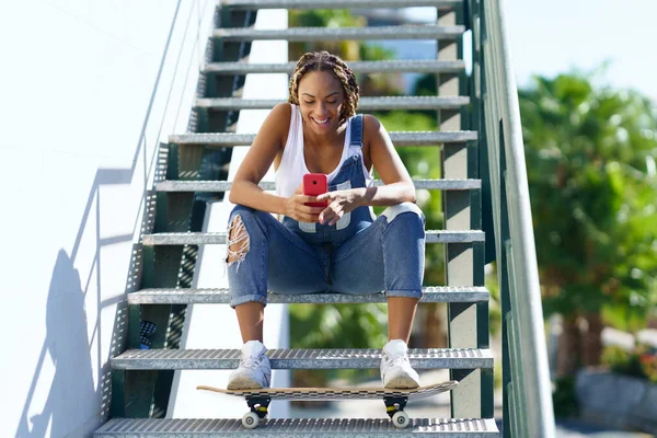 Schwarze Frau mit farbigen Zöpfen, die mit ihren Füßen auf einem Skateboard ihr Smartphone konsultiert. — Stockfoto