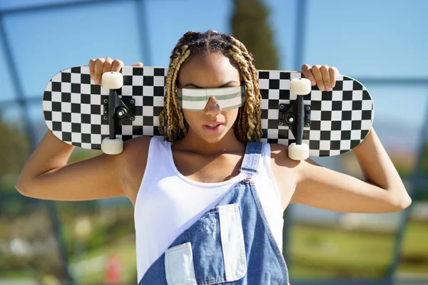 Черная женщина одета непринужденно, смотрит в камеру в современных солнцезащитных очках и скейтборде. — стоковое фото