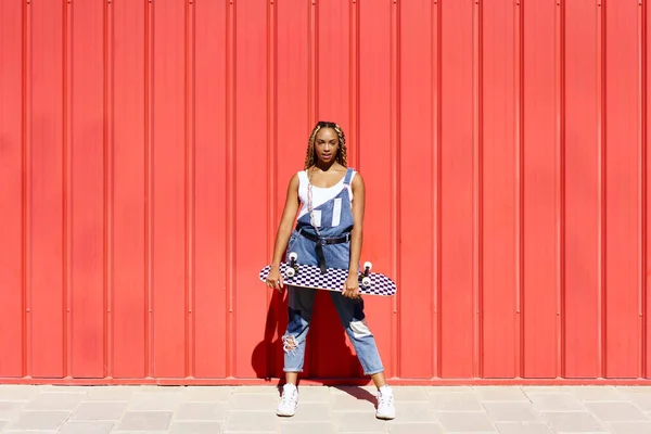 Черная женщина одета непринужденно, скейтборд на фоне красной городской стены. — стоковое фото