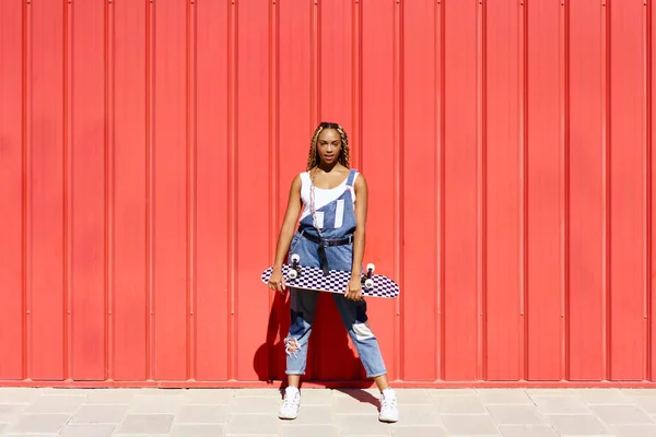 Mulher negra vestida casual, wtih um skate no fundo da parede urbana vermelha. — Fotografia de Stock