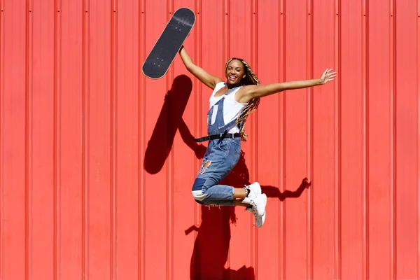 Donna nera vestita casual, wtih uno skateboard che salta con felicità su sfondo muro urbano rosso. — Foto Stock