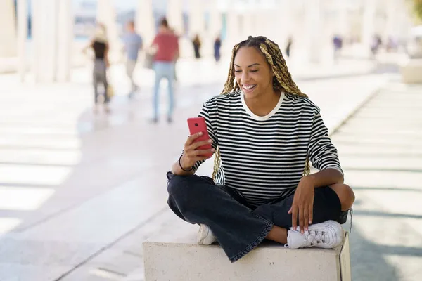 Gelukkig zwart vrouw met behulp van een smartphone buiten, het dragen van haar haar in vlechten. — Stockfoto