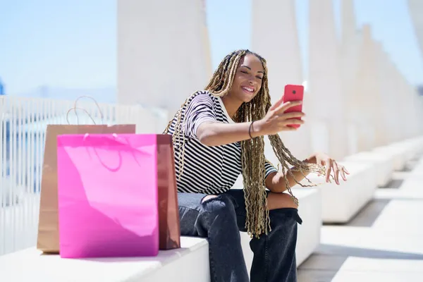 Siyah bir kadın akıllı telefonuyla selfie çekiyor. Sokakta bir bankta oturuyor.. — Stok fotoğraf