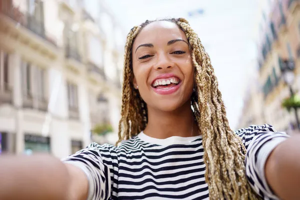 Zwarte vrouw met afro vlechten nemen van een lachende selfie in een stedelijke straat met een smartphone. — Stockfoto