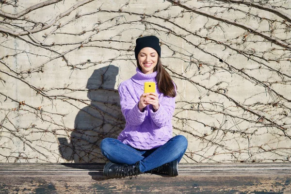 Boldog fiatal nő mosolyog ül a padon a szabadban egy okostelefon segítségével. — Stock Fotó