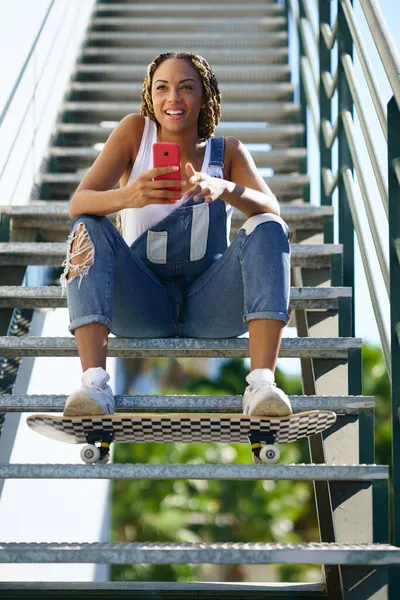 Czarna dziewczyna z kolorowymi warkoczami, siedząca ze smartfonem i deskorolką. — Zdjęcie stockowe