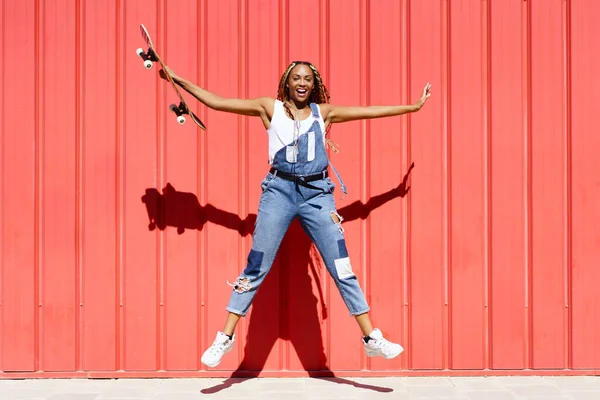 Mujer negra vestida casual, con un monopatín saltando para joyon fondo de pared urbana roja. — Foto de Stock