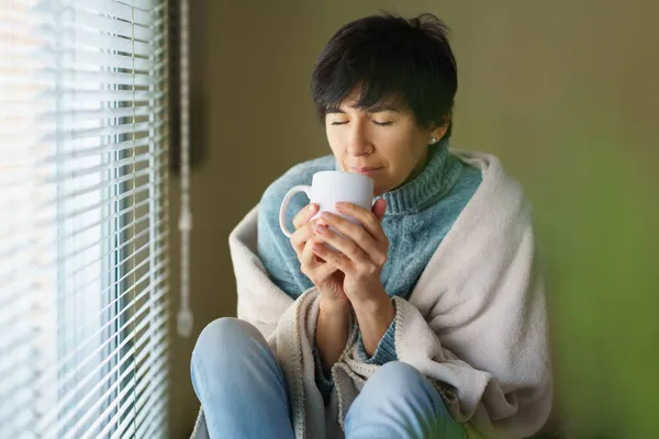 Donna di mezza età che sente odore di caffè dalla sua tazza vicino a una finestra. — Foto Stock