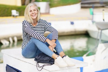 Gülümseyen yaşlı kadın kameraya sakince bakıyor, bir limanda oturuyor..