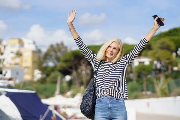Woman in her 60s strolling along a seaside spot near the beach, raising her arms in joy. — Stock Photo, Image