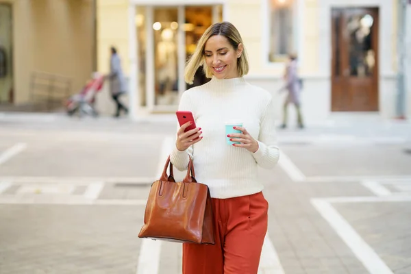 Mujer caminando por la calle usando un teléfono inteligente mientras bebe café caliente en un vaso para llevar. — Foto de Stock