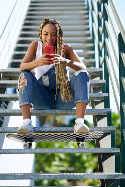 Zwarte vrouw, met gekleurde vlechten, kijken naar iets grappigs op haar smartphone. — Stockfoto