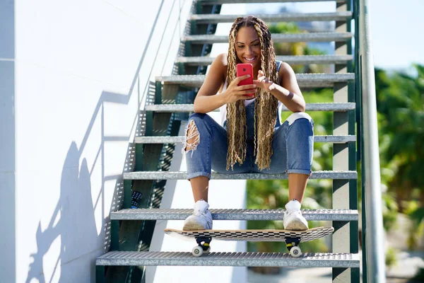 Schwarze Frau mit farbigen Zöpfen, die mit ihren Füßen auf einem Skateboard ihr Smartphone konsultiert. — Stockfoto