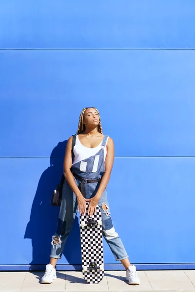 Black girl dressed casual, wtih a skateboard on blue wall background. — 图库照片