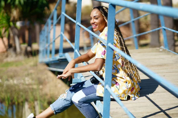 身穿辫子的非洲女孩坐在一座城市桥上度假时拿着照相机 — 图库照片
