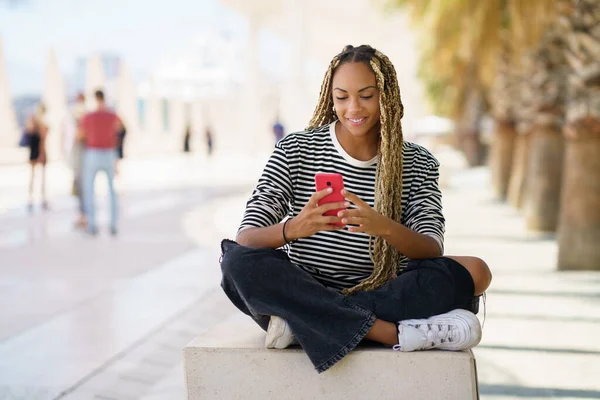 Zwarte vrouw sms 'en met een smartphone zitten op een bank buiten, het dragen van haar haar in vlechten. — Stockfoto