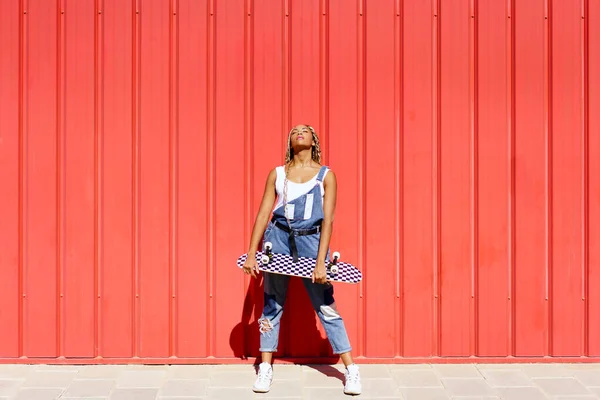 Chica negra vestida casual, con un monopatín sobre fondo rojo de pared urbana. —  Fotos de Stock