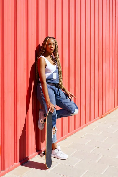 黒の女性がカジュアルな服装,赤い都市の壁の背景にスケートボードと. — ストック写真