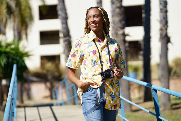 Glad svart kvinna kammad med färgglada flätor promenader med en kamera på semester. — Stockfoto