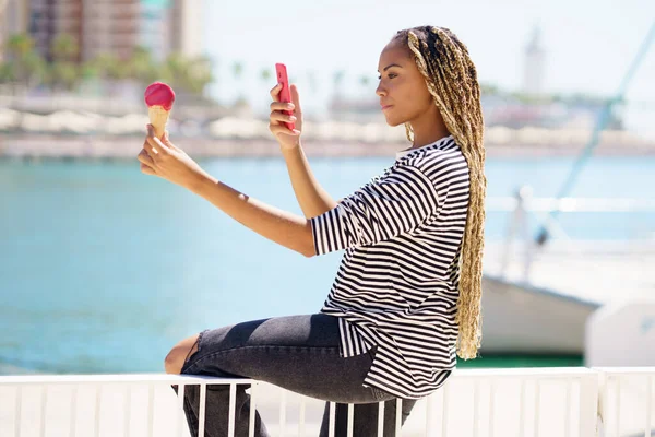 Black girl photographing with her smartphone a strawberry ice cream she is eating near the sea — Foto Stock