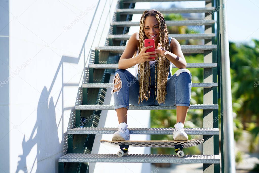Black woman with coloured braids, consulting her smartphone with her feet resting on a skateboard.