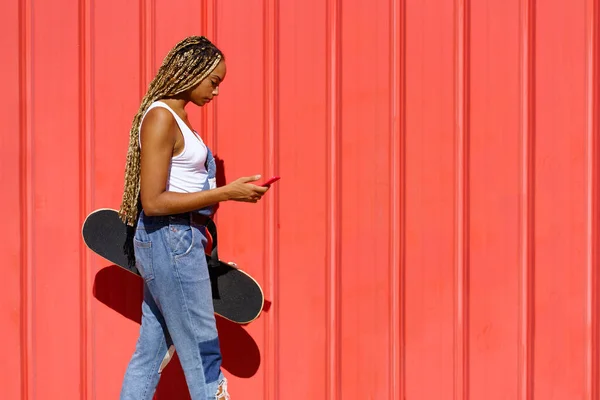 Zwart meisje lopen op een stedelijke rode achtergrond tijdens het raadplegen van haar smartphone. — Stockfoto