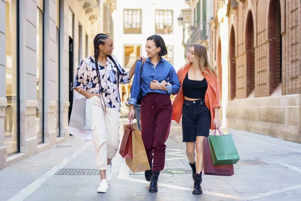 Stílusos multiracionális női bolti mániákusok sétálnak együtt az utcán papírzacskókkal — Stock Fotó