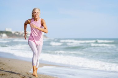 Yaşlı kadın formda kalmak için spor yapıyor. Kumsalda koşan olgun bir kadın..