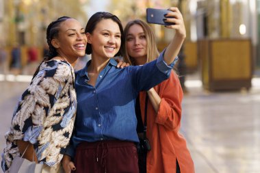 Akıllı telefondan selfie çekerken gülümseyen çok ırklı genç bayan turistler.