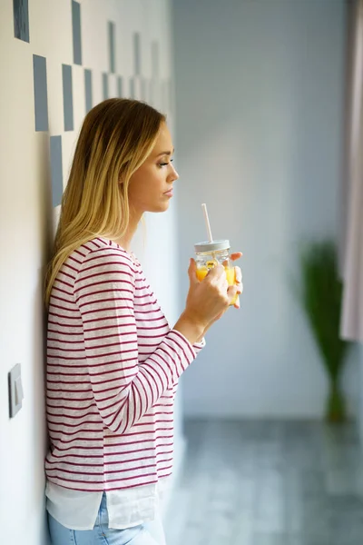 Mujer pensativa bebiendo un vaso de jugo de naranja natural en casa. — Foto de Stock