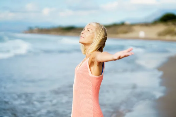 Piękna starsza kobieta rozkłada ramiona na pięknej plaży, spędzając wolny czas. — Zdjęcie stockowe