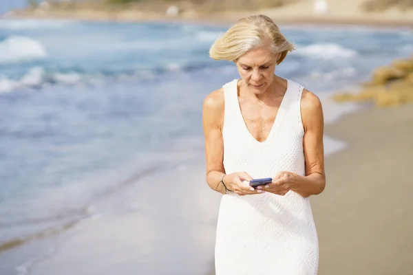 Senioren die met een smartphone over het strand lopen. — Stockfoto