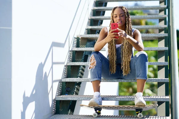 Mujer negra con trenzas de colores, consultando su smartphone con los pies apoyados en un monopatín. —  Fotos de Stock