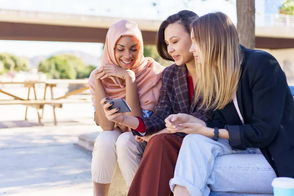 Positive multiethnische Freundinnen teilen Smartphone, während sie sich auf Bank im Park ausruhen — Stockfoto