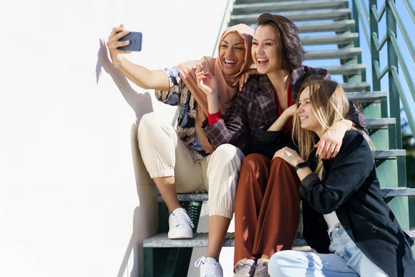 Felice multietnico femminile migliori amici prendendo selfie seduto sulle scale in — Foto Stock