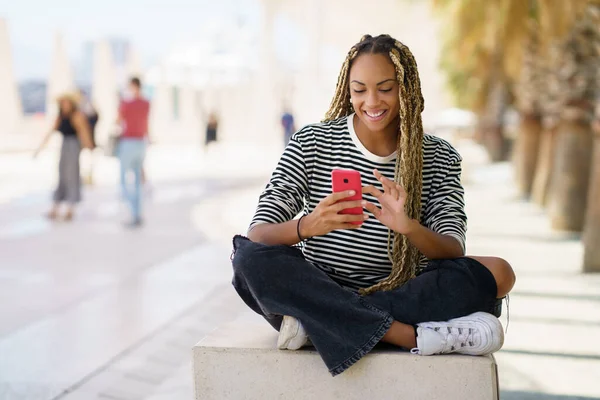 Zwart meisje sms 'en met een smartphone zitten op een bank buiten, het dragen van haar haar in vlechten. — Stockfoto