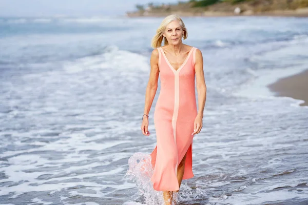 Senior vrouw in jurk wandelen langs het strand — Stockfoto